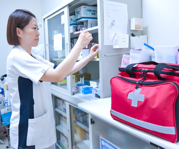 ナースステーションや薬品庫などの棚の前で、女性スタッフが医療資材や医薬品の確認・管理を行っている様子（手前に赤い救急バッグ）