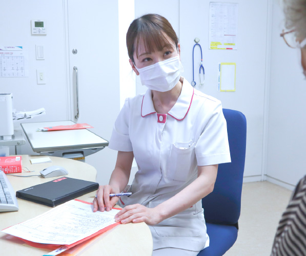 診察室（または相談室）にて、マスクを着用した白いユニフォームの女性スタッフが机を挟んで患者（または家族）の相談に熱心に耳を傾けている様子
