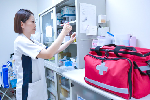 ナースステーションや薬品庫などの棚の前で、女性スタッフが医療資材や医薬品の確認・管理を行っている様子（手前に赤い救急バッグ）