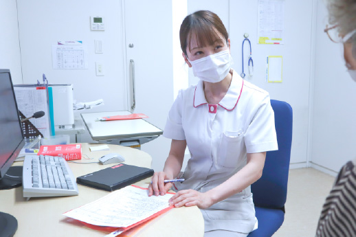 診察室（または相談室）にて、マスクを着用した白いユニフォームの女性スタッフが机を挟んで患者（または家族）の相談に熱心に耳を傾けている様子