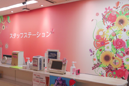 ひまわりなどの華やかなイラストが描かれたピンク色の壁面が特徴的な、明るいスタッフステーション（ナースステーション）の受付カウンターの様子