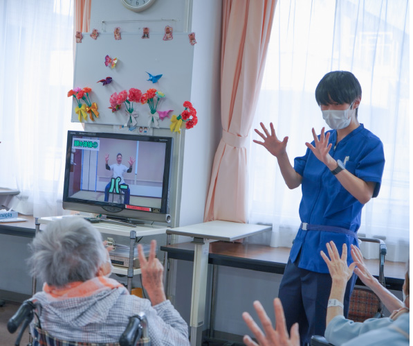 高齢者施設で、職員がモニターを使った体操を利用者と一緒に行っている様子
