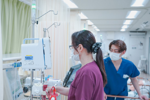 透析室にて、医療スタッフが透析機器の前に立ち、機器の操作や管理・確認を行っている様子