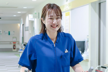病院の廊下で、青いスクラブを着用した女性スタッフが笑顔で歩行介助（または移動のサポート）をしている様子