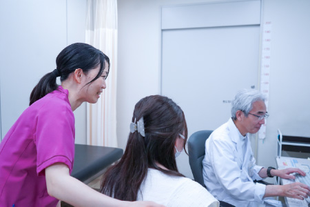 診察室にて、白衣の医師がパソコンに向かう傍らで、ピンクのスクラブを着用した女性スタッフが患者に寄り添いサポートしている様子