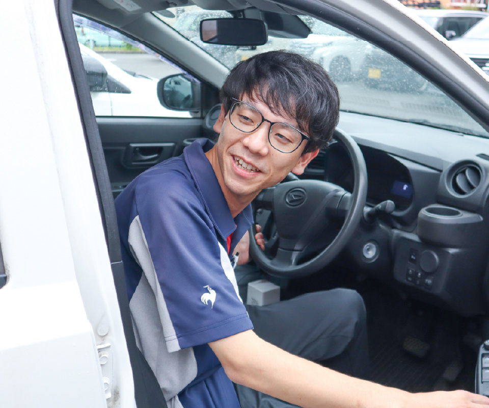 紺色のポロシャツを着用した男性スタッフが、訪問看護のために車の運転席に乗り込み、笑顔で出発する様子