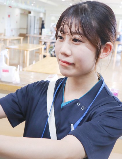 紺色のスクラブを着用した女性スタッフが、明るく広々とした食堂（またはデイルーム）のような場所で、手元に意識を向けながら優しい表情で行対している様子