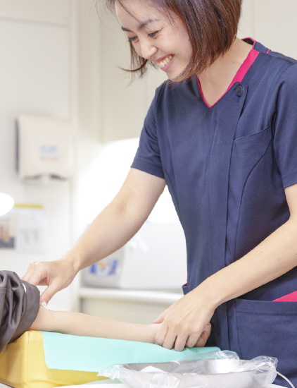 紺色のスクラブを着用した女性スタッフが、笑顔で患者の腕（または手元）に優しく触れながらケアや処置を行っている様子