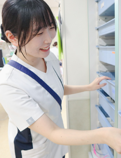 白いユニフォームを着用した女性スタッフが、笑顔でカルテ棚や薬品棚から書類・資材を取り出している様子