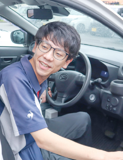 紺色のポロシャツを着用した男性スタッフが、車の運転席に乗り込み、笑顔で出発する様子